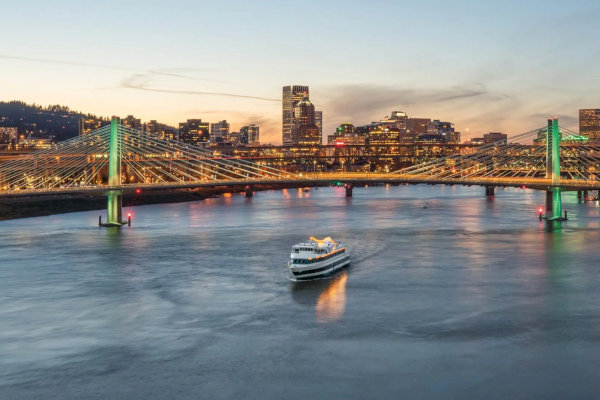 The image shows a city skyline at dusk with a brightly lit bridge spanning a river, where a boat is cruising towards the shore.