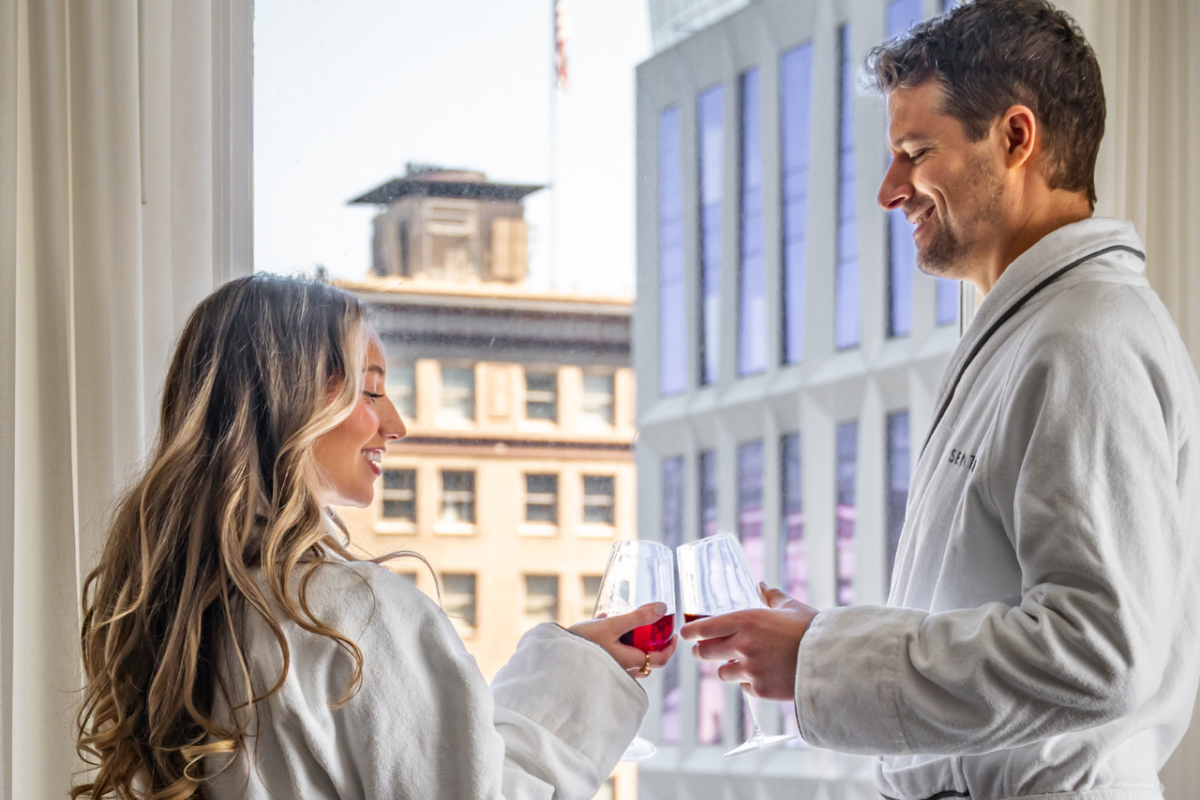 A man and a woman in bathrobes smile at each other while holding glasses of red wine inside a building with a view of the city outside the window.
