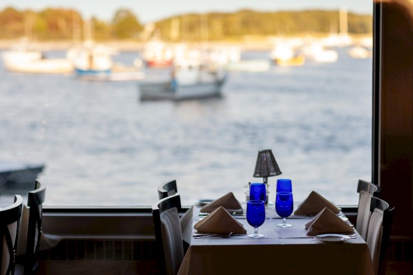 A set dinner table by a window overlooks a scenic waterfront with boats floating on the water in the background, creating a serene view.