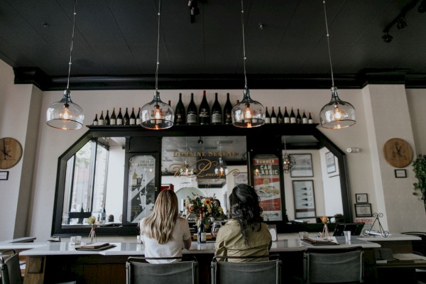 Two people sitting at a bar with bottles displayed above and hanging lights illuminating the area. The sentence ends.
