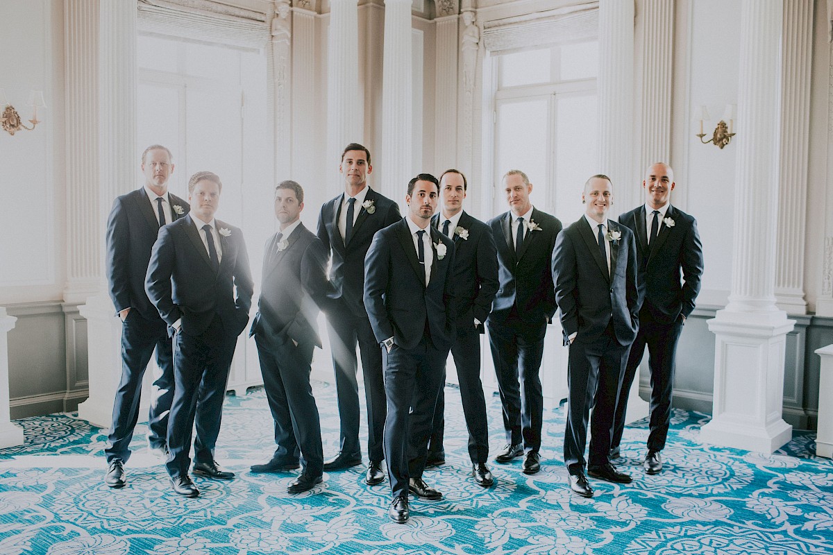 A group of nine men dressed in formal suits posing indoors with bright natural light; the setting has elegant decor and blue patterned carpet.