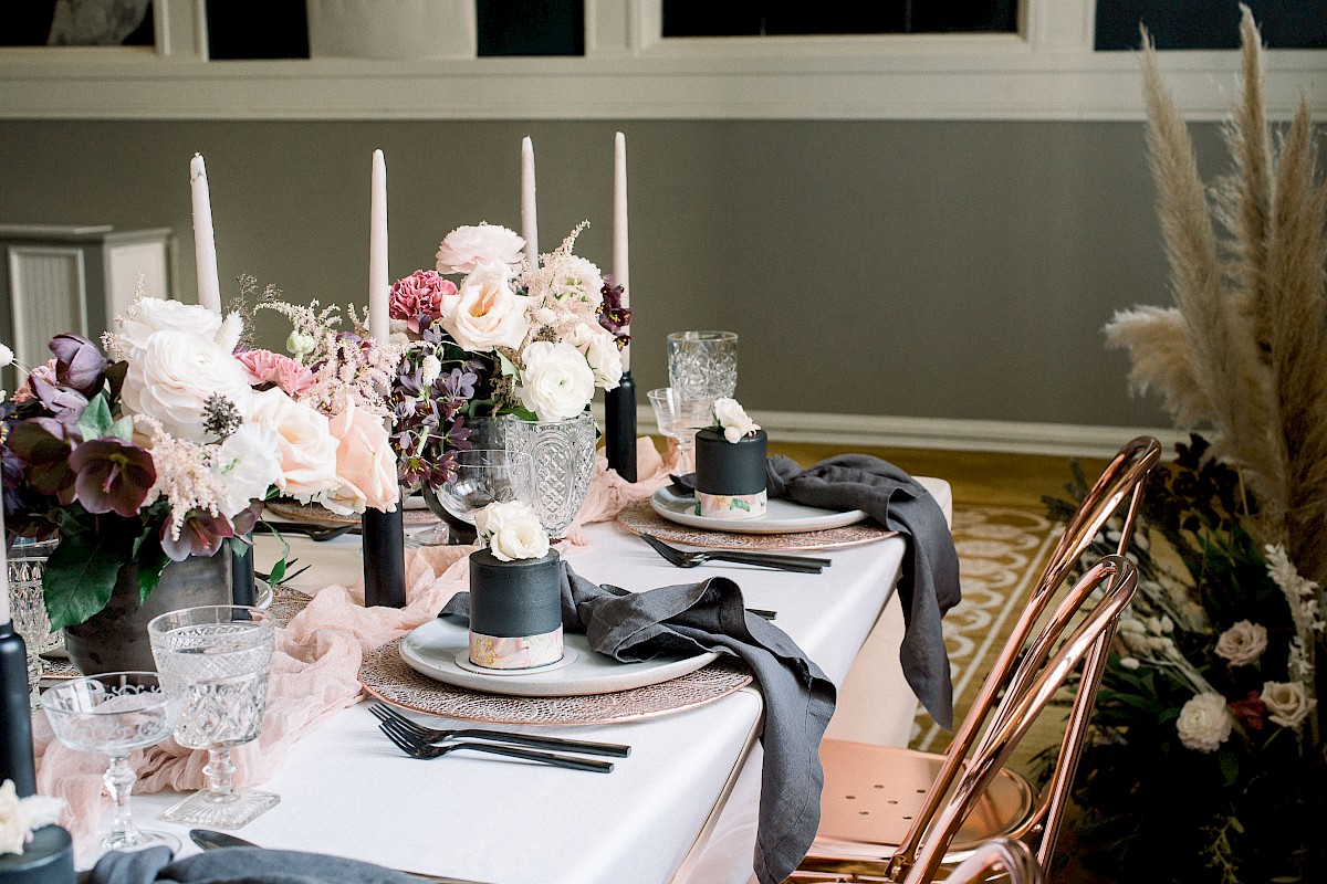 A beautifully set dining table with flowers, candles, glassware, and silverware arranged elegantly, featuring rose-gold chairs and gray napkins.