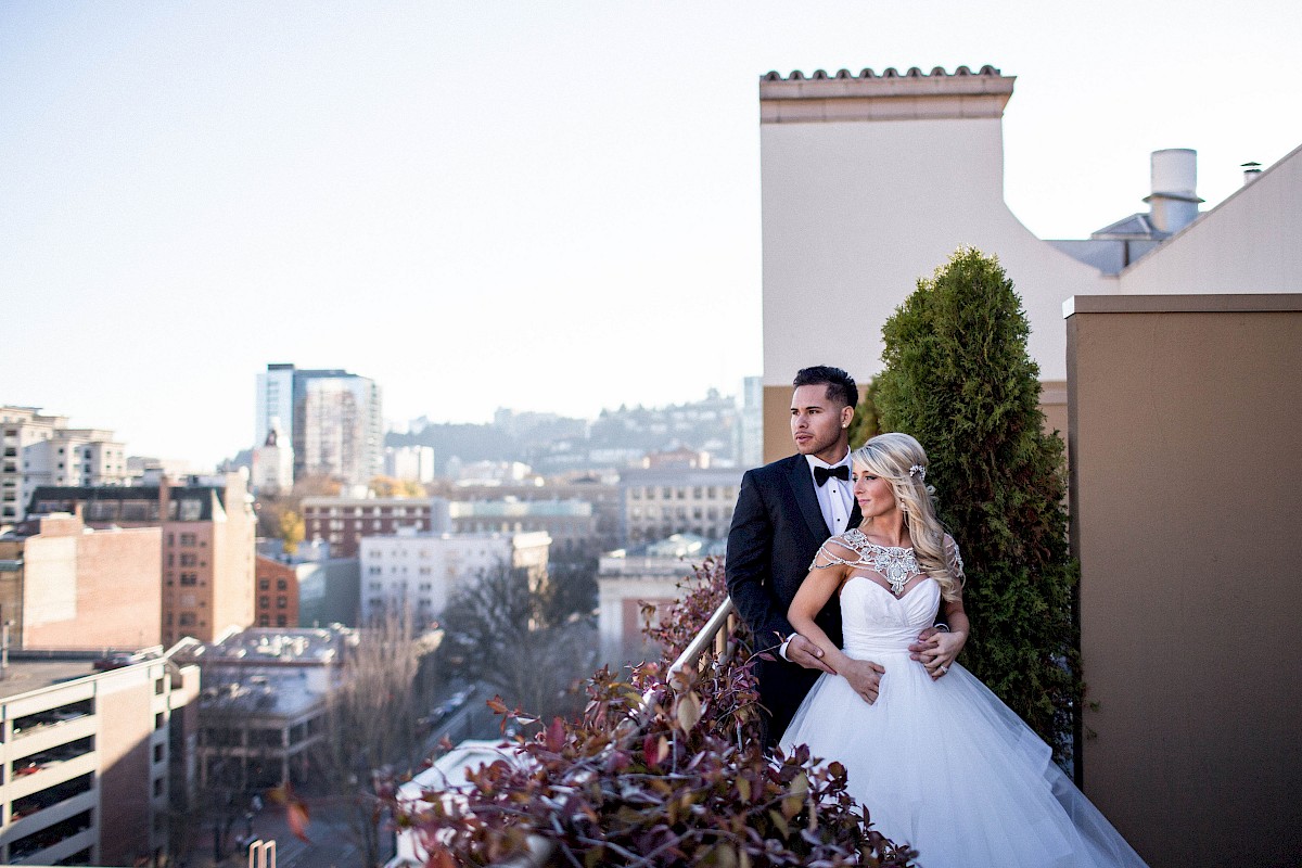 A couple in formal wedding attire poses on a rooftop with a cityscape in the background; the groom is in a tuxedo, and the bride is in a white gown.