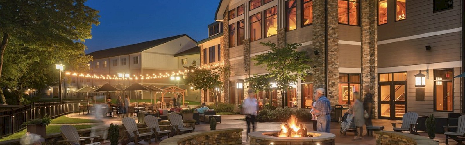 An evening scene of a large house with a lit fire pit as people lounge in chairs. String lights are hanging between buildings, creating a cozy atmosphere.