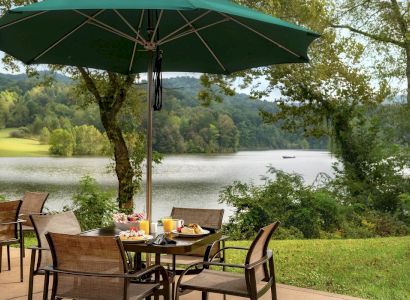 An outdoor dining area with a table set for a meal under a large green umbrella, overlooking a scenic lake surrounded by trees and hills.