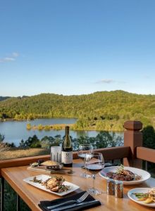 A table with food, wine, and utensils set on a balcony overlooking a scenic lake and forested hills under a clear blue sky.