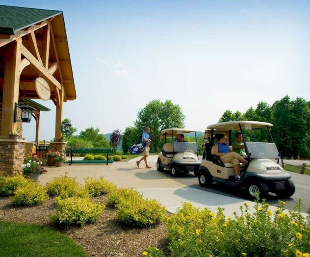 The image shows a golf course with two people in golf carts and a person carrying golf clubs walking towards them. Bright, sunny day outdoors.