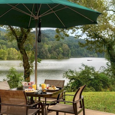An outdoor dining area features a table with food and drinks under a large green umbrella, overlooking a serene lake and lush, tree-covered hills.