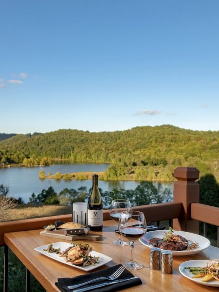 A wooden table with a meal, wine, and glasses is set on a deck overlooking a serene lake and lush green hills under a clear blue sky.