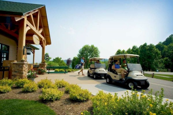 Two people are driving golf carts near a building with wood accents. Another person walks away carrying a golf bag. There's greenery around.