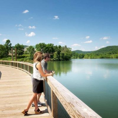 Two people stand on a wooden boardwalk, overlooking a scenic lake surrounded by trees and hills under a clear blue sky with scattered clouds.