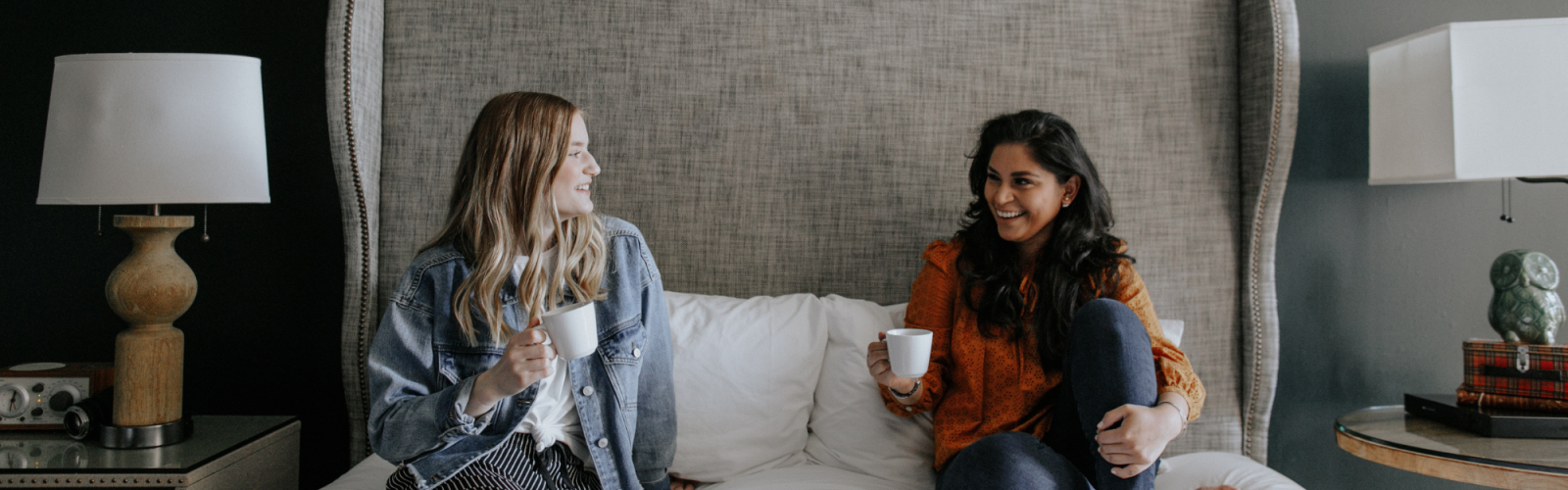 Two women sit on a bed, enjoying drinks and a breakfast spread, smiling at each other.