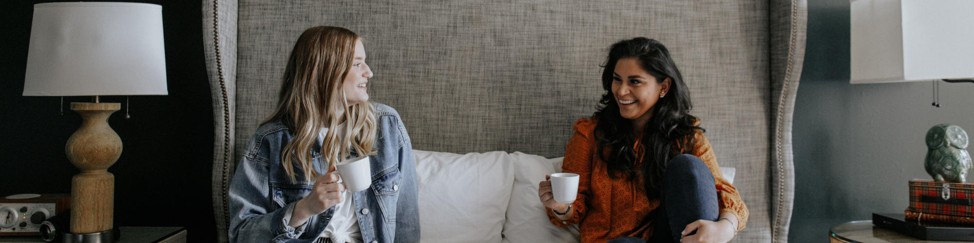 Two women sit on a bed, enjoying drinks and a breakfast spread, smiling at each other.