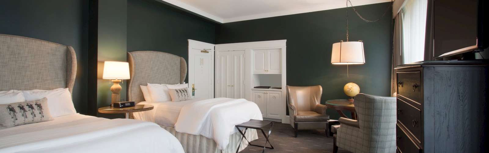 A hotel room with two queen beds, nightstands with lamps, a TV, chairs, a globe, and a cabinet, featuring dark green walls and carpeted flooring.