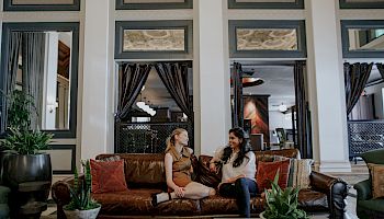 Two women sitting and chatting on a brown sofa in a stylish, plant-decorated lounge area with large windows and neutral tones ending the sentence.