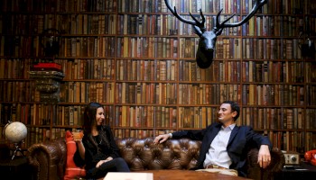 A man and woman sit on a leather couch in a library with a wall of books, a globe, and a mounted deer head, engaged in conversation.