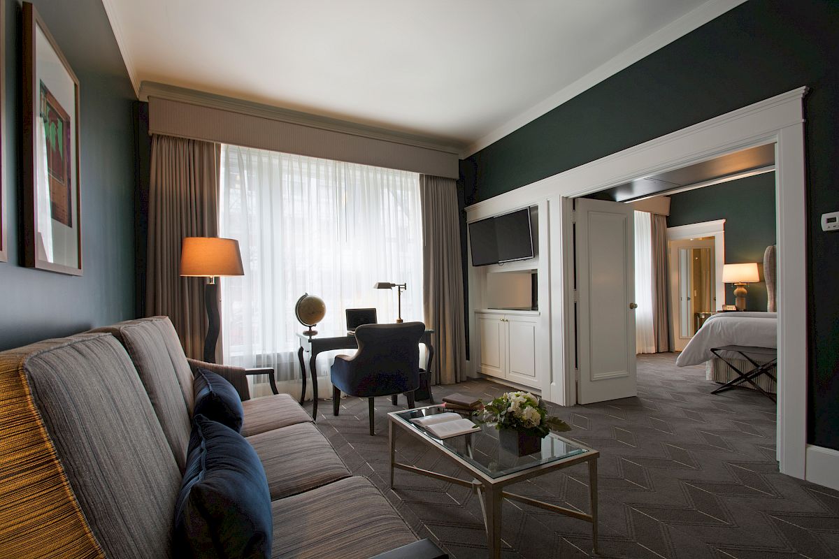 A modern hotel room with a seating area, desk, sofa, lamp, and coffee table, leading into a bedroom through open double doors.