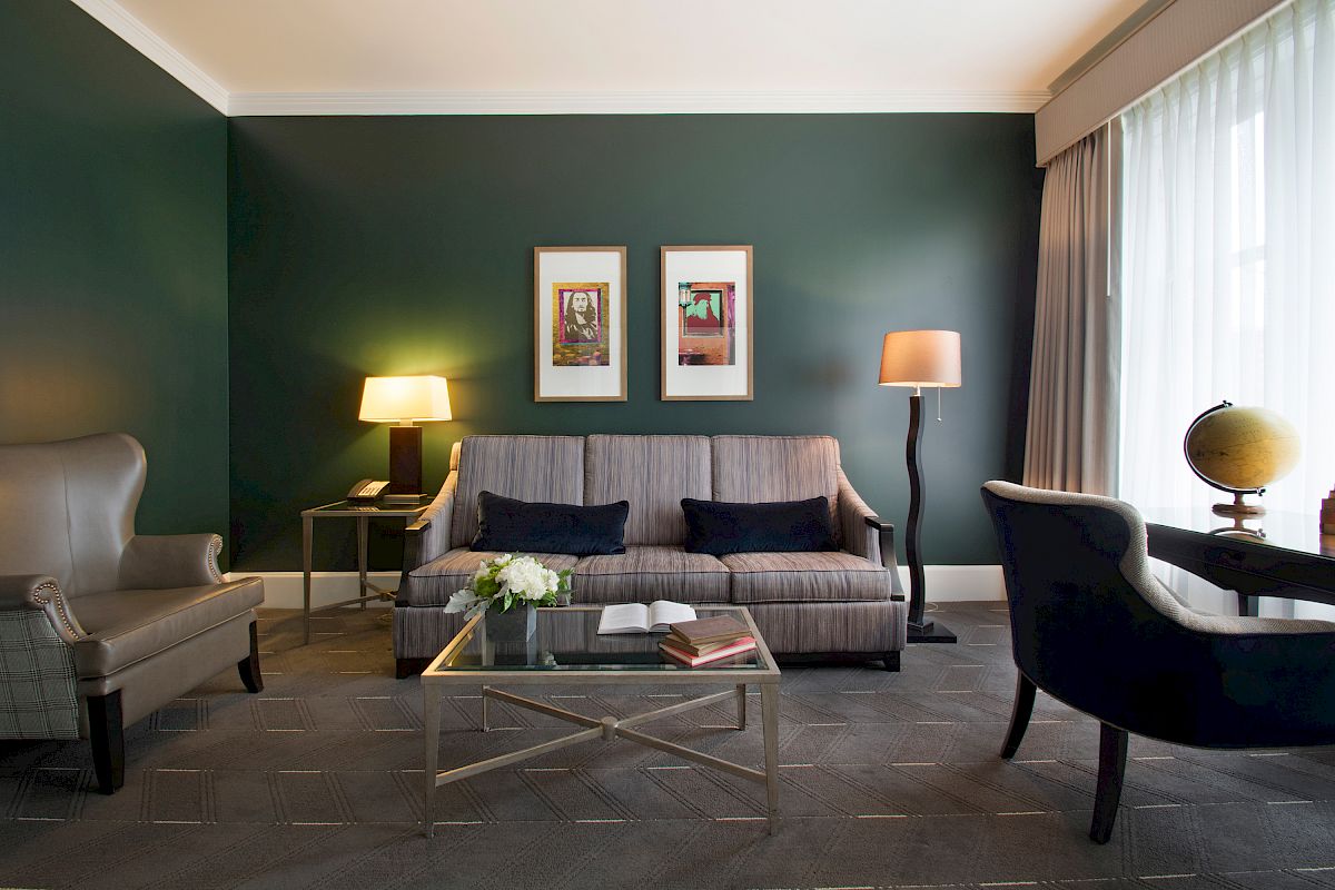 A modern living room with a sofa, two armchairs, two lamps, a glass coffee table, framed artwork, and a desk with a globe near a large window with curtains.