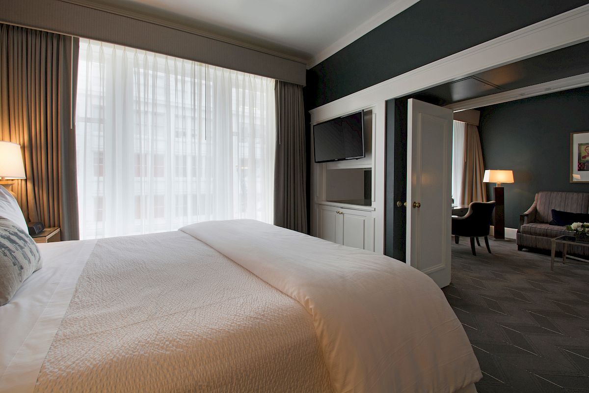 This image shows a hotel room with a large bed, a TV on the wall, and a seating area in an adjoining room with large windows and curtains.