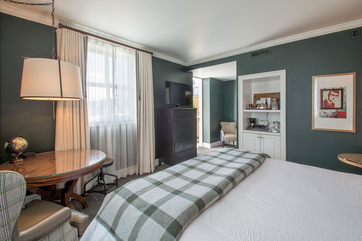 A cozy bedroom with green walls, a checkered blanket on the bed, a round table, a TV on a dresser, and artwork on the wall.