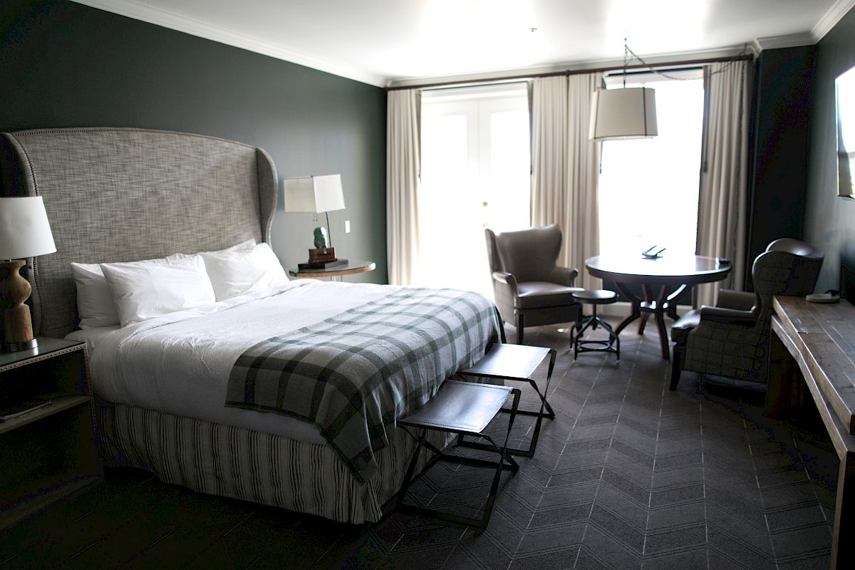 A stylish hotel room features a large bed with a gray headboard, a small table with chairs, a desk, and large windows with curtains.