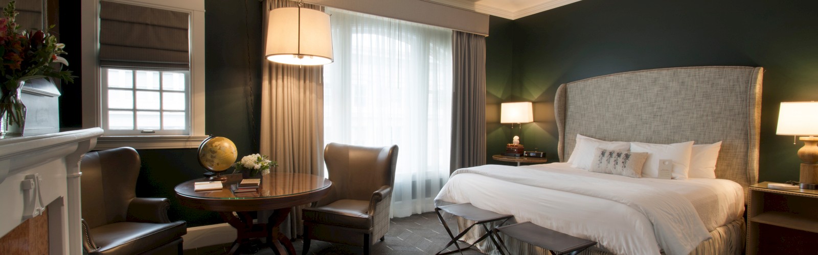 A cozy hotel room features a large bed, two armchairs, a small table, a lamp, and a fireplace. The room has dark green walls and a large window.