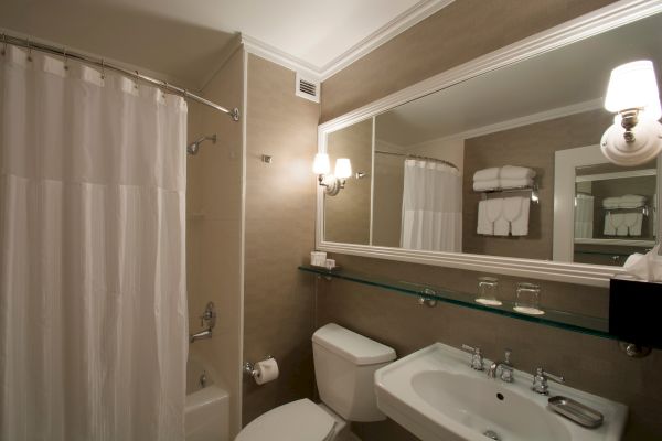 A clean, well-lit bathroom with a shower curtain, toilet, sink, large mirror, wall-mounted light, and shelves with towels and glasses.