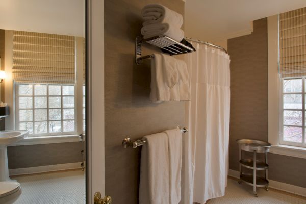 This image shows a bathroom with a pedestal sink, shower with a curtain, towels on a rack, two windows with blinds, and a small shelf.