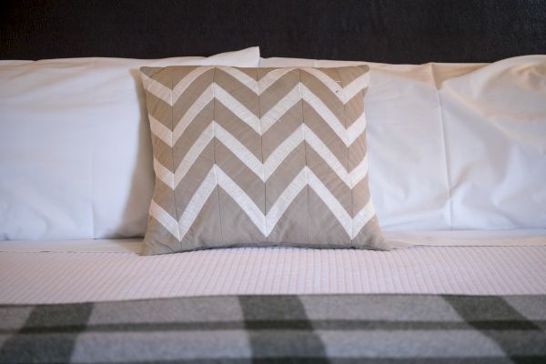 A bed with white pillows and a beige chevron-patterned cushion in the center, topped with a grey and white checkered blanket.