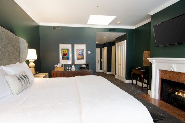 A cozy hotel room features a large bed, wall art, a fireplace, TV, and a small refreshment station with snacks and drinks.