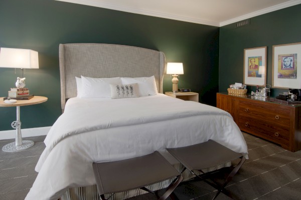 A stylish bedroom with a large bed, bedside lamps, art on the wall, a wooden dresser, and a basket.