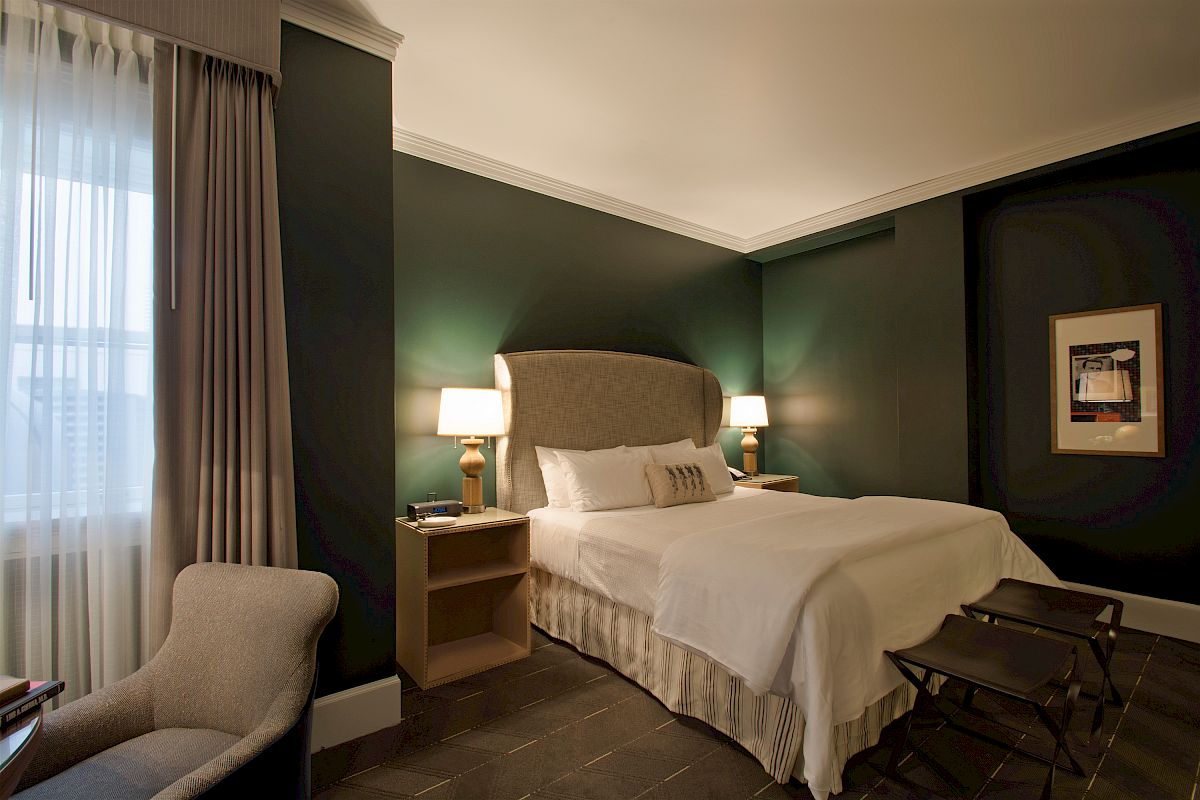 A cozy hotel room with a large bed, two bedside tables with lamps, a window with curtains, an armchair, and framed artwork on the wall, always ending the sentence.