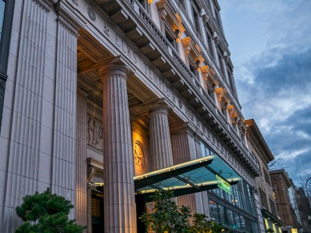 The image shows the exterior of a grand, lighted building with tall columns and ornate designs. Trees are placed near the entrance.