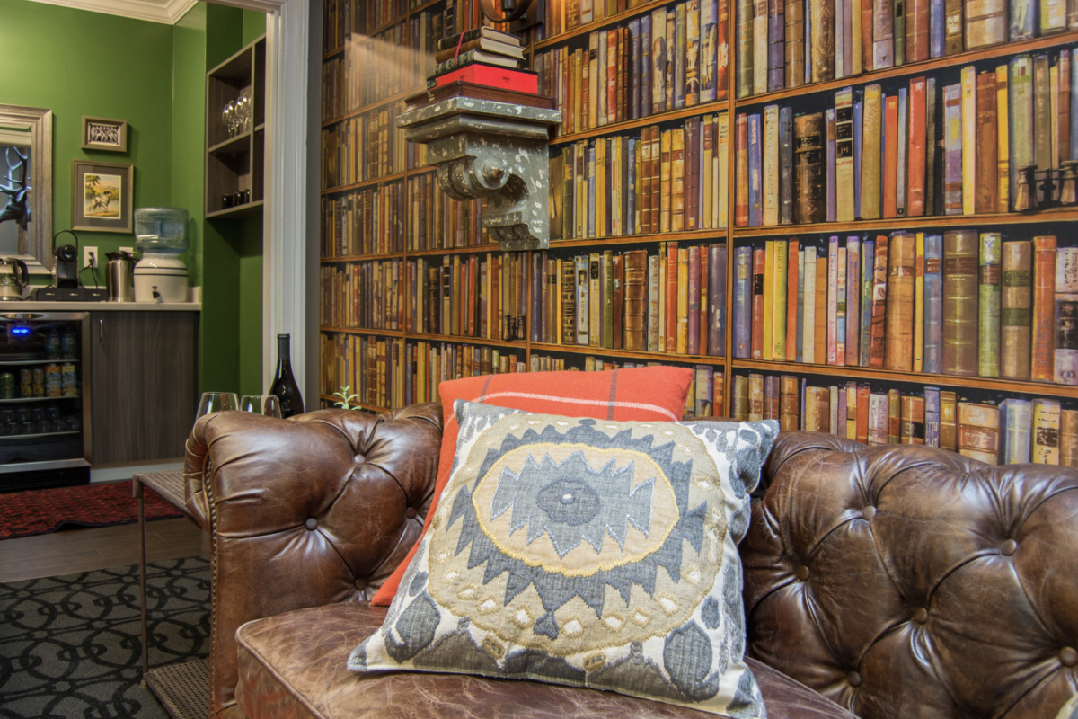 A cozy room with a brown leather couch, a decorative pillow, a bookshelf wallpaper, and a kitchenette in the background featuring a water dispenser.