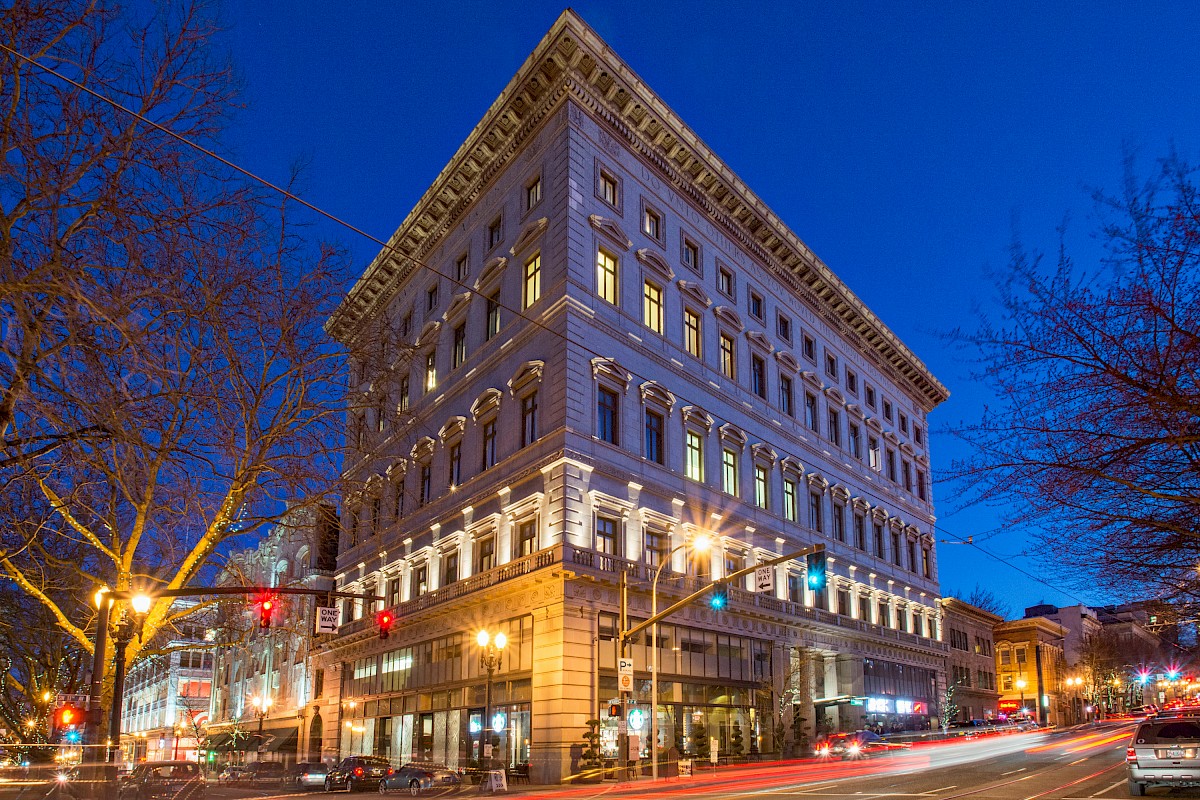 A well-lit, historic multi-story building on a street corner at dusk, with light trails from passing vehicles and bare trees nearby.