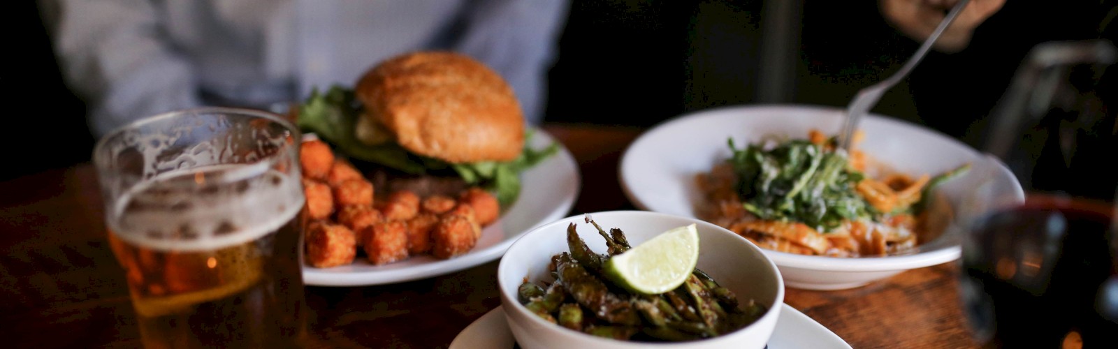The image shows a wooden table with a bowl of food, a burger with tater tots, a glass of beer, another plate being eaten, and a glass of red wine.