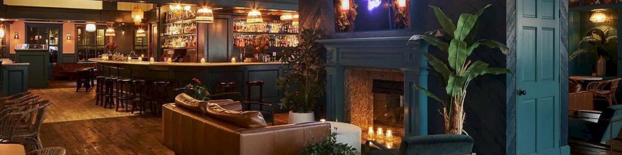 The image shows a cozy, modern bar with wooden floors, a fireplace, a bar area, and lounge seating, featuring warm lighting and contemporary decor.
