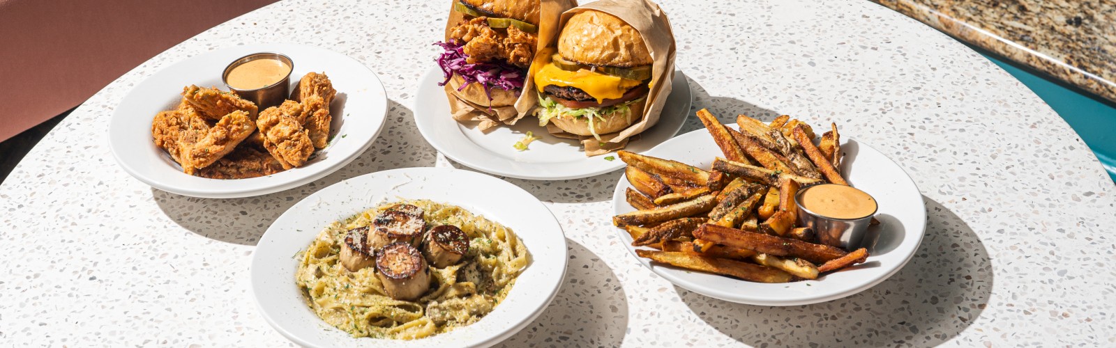 The image shows a table with various dishes: burgers, fries, pasta with mushrooms, and chicken with dipping sauce, against a cozy interior setting.