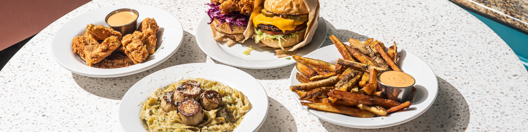 The image shows a table with various dishes: burgers, fries, pasta with mushrooms, and chicken with dipping sauce, against a cozy interior setting.