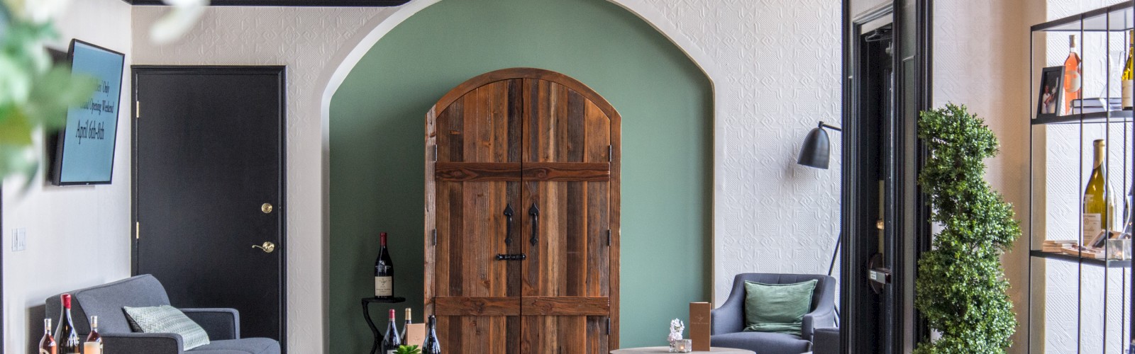 The image shows a modern sitting area with wooden tables, green chairs, a wooden door in an archway, and various bottles on display shelves.