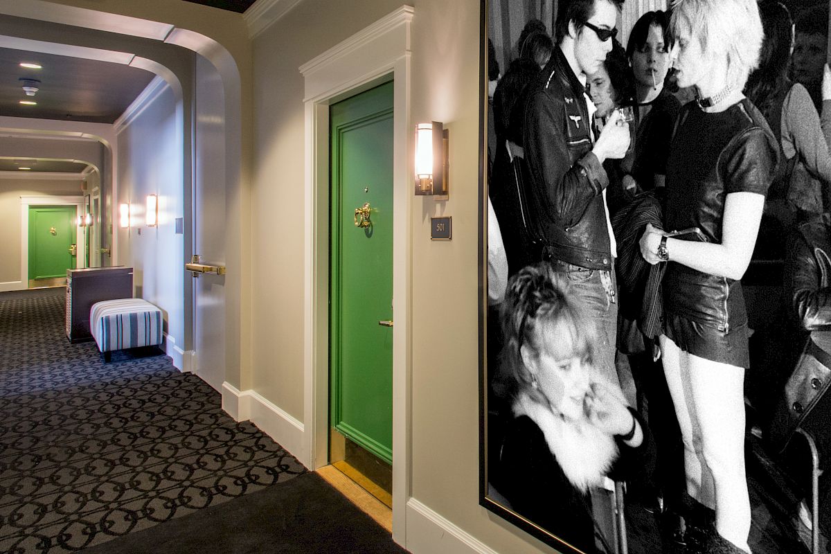 A hotel hallway with green doors and patterned carpet, featuring a large black-and-white photo of people socializing on the wall.