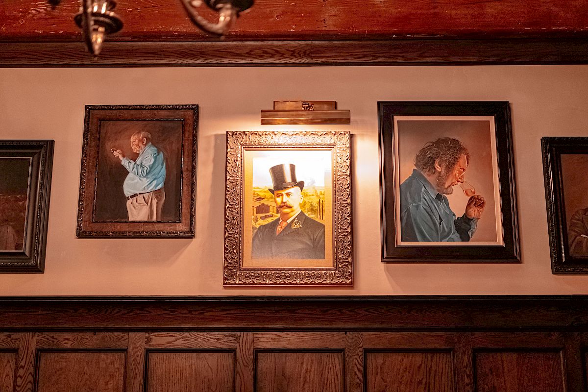 Three framed portraits are displayed on a wooden wall; two depict men in blue attire, and the center portrait is of a man with a top hat.
