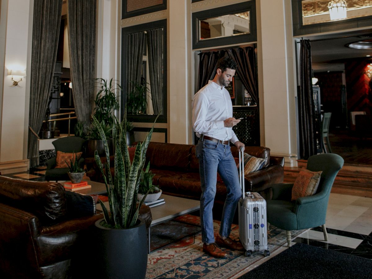 A person stands in a luxurious room with ornate ceilings, holding a suitcase near elegant furniture and indoor plants.