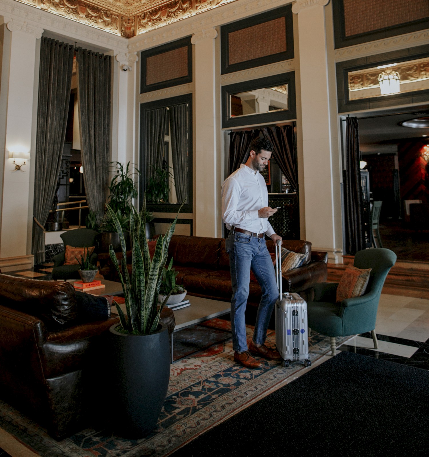 A person stands in a luxurious room with ornate ceilings, holding a suitcase near elegant furniture and indoor plants.