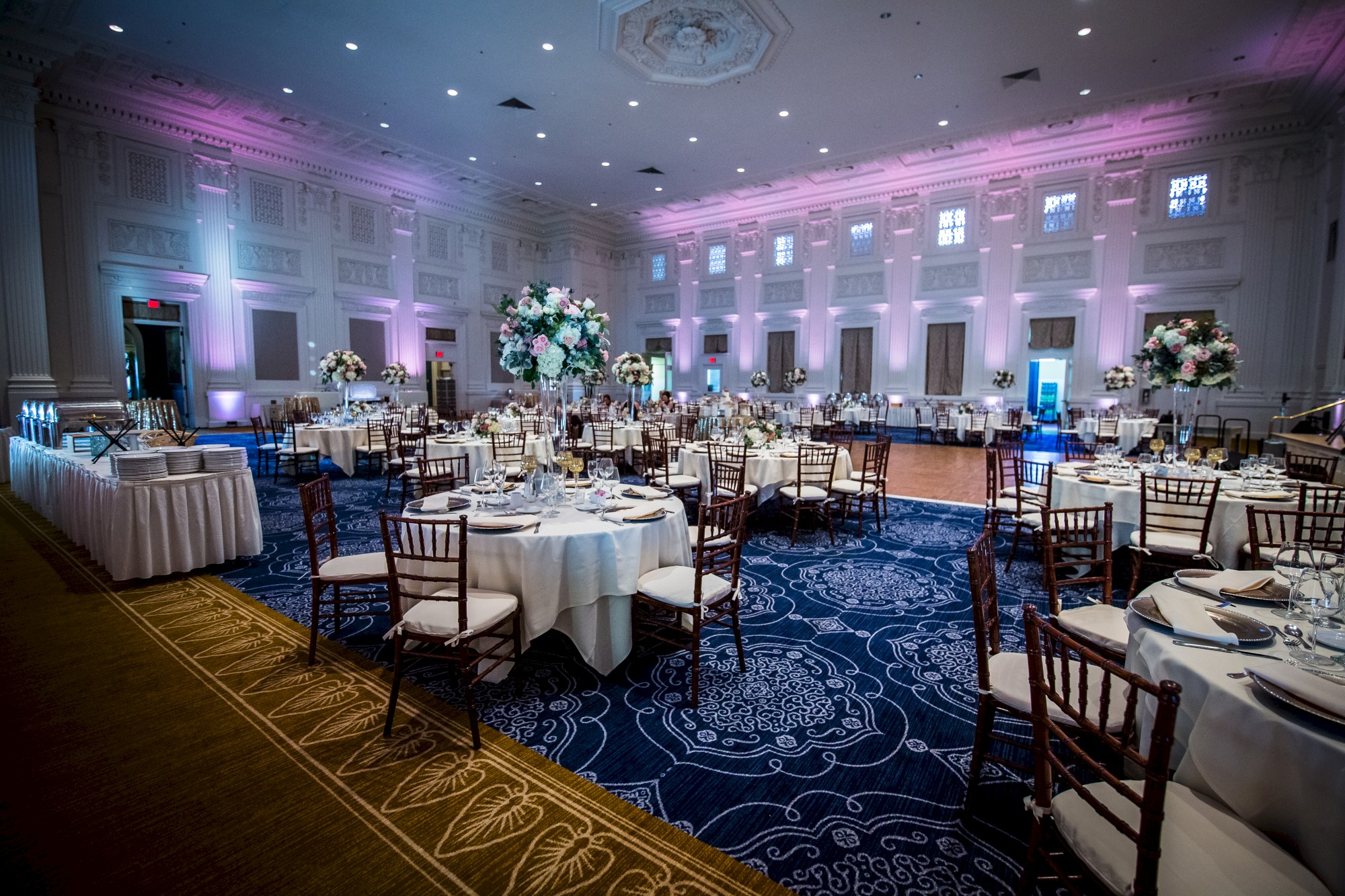 A large, elegantly decorated banquet hall with round tables, floral centerpieces, and purple lighting. Tables are set for a formal event.