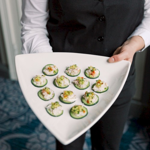 A person in a black vest and white shirt holds a triangular plate with bite-sized appetizers, likely on cucumber slices with various toppings.