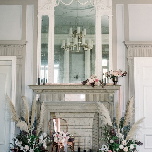 An elegant fireplace decorated with flowers and pampas grass arrangements. A large mirror above reflects a chandelier, enhancing the room’s grandeur.