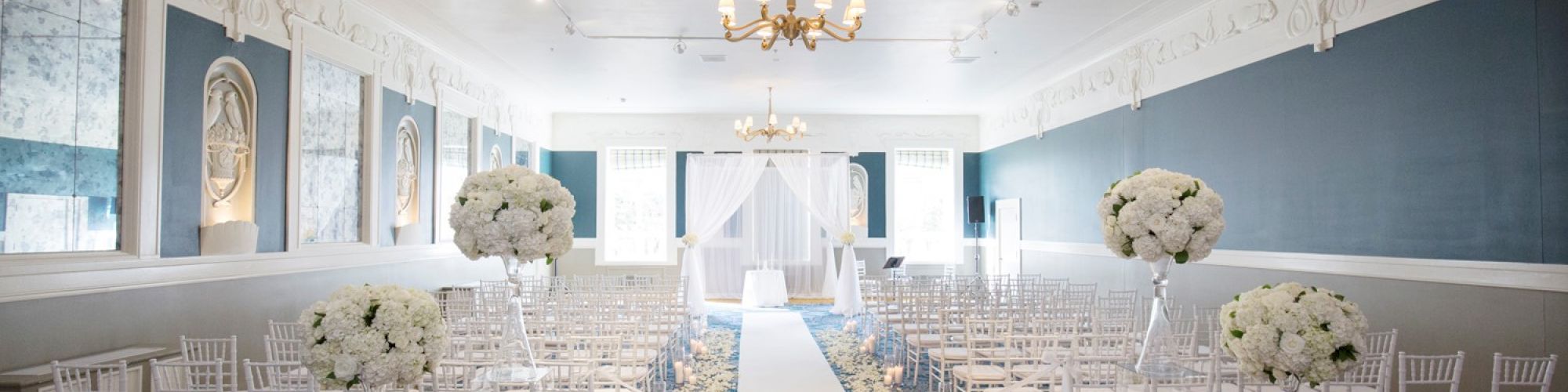 A wedding ceremony setup with rows of white chairs, floral arrangements, and a white aisle runner in an elegant, well-lit room ending the sentence.