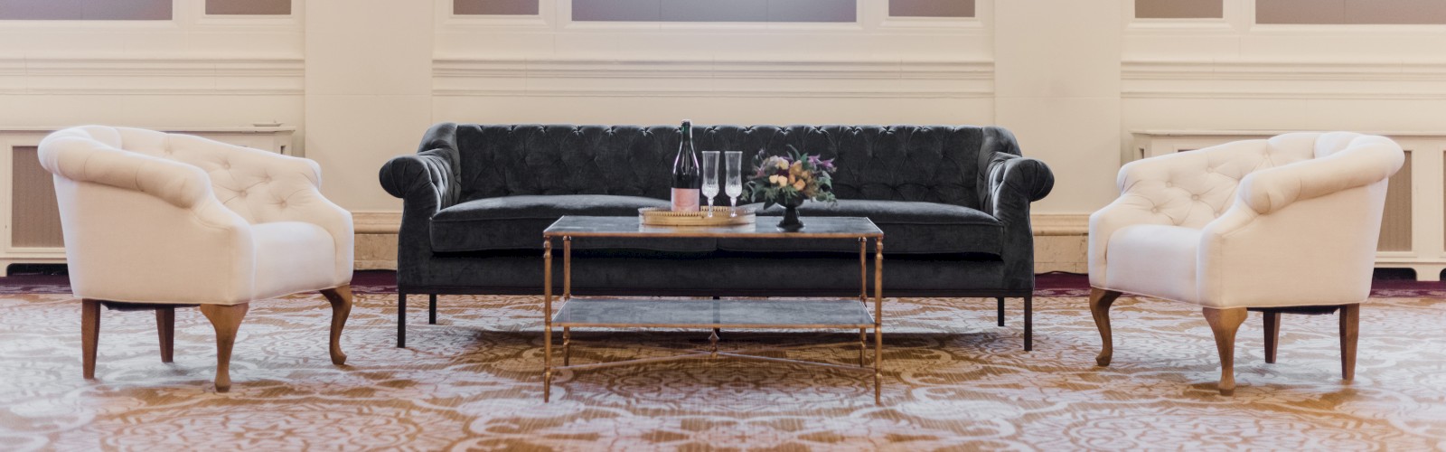 A sophisticated seating area with two white chairs, a black sofa, a coffee table, and a champagne bottle in a room with tall windows and curtains.