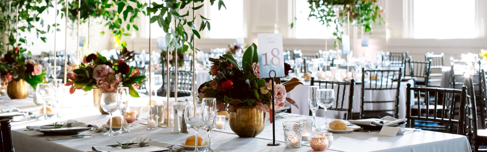 A beautifully decorated banquet hall with elegantly set tables, lush floral centerpieces, and tall windows allowing natural light to pour in.